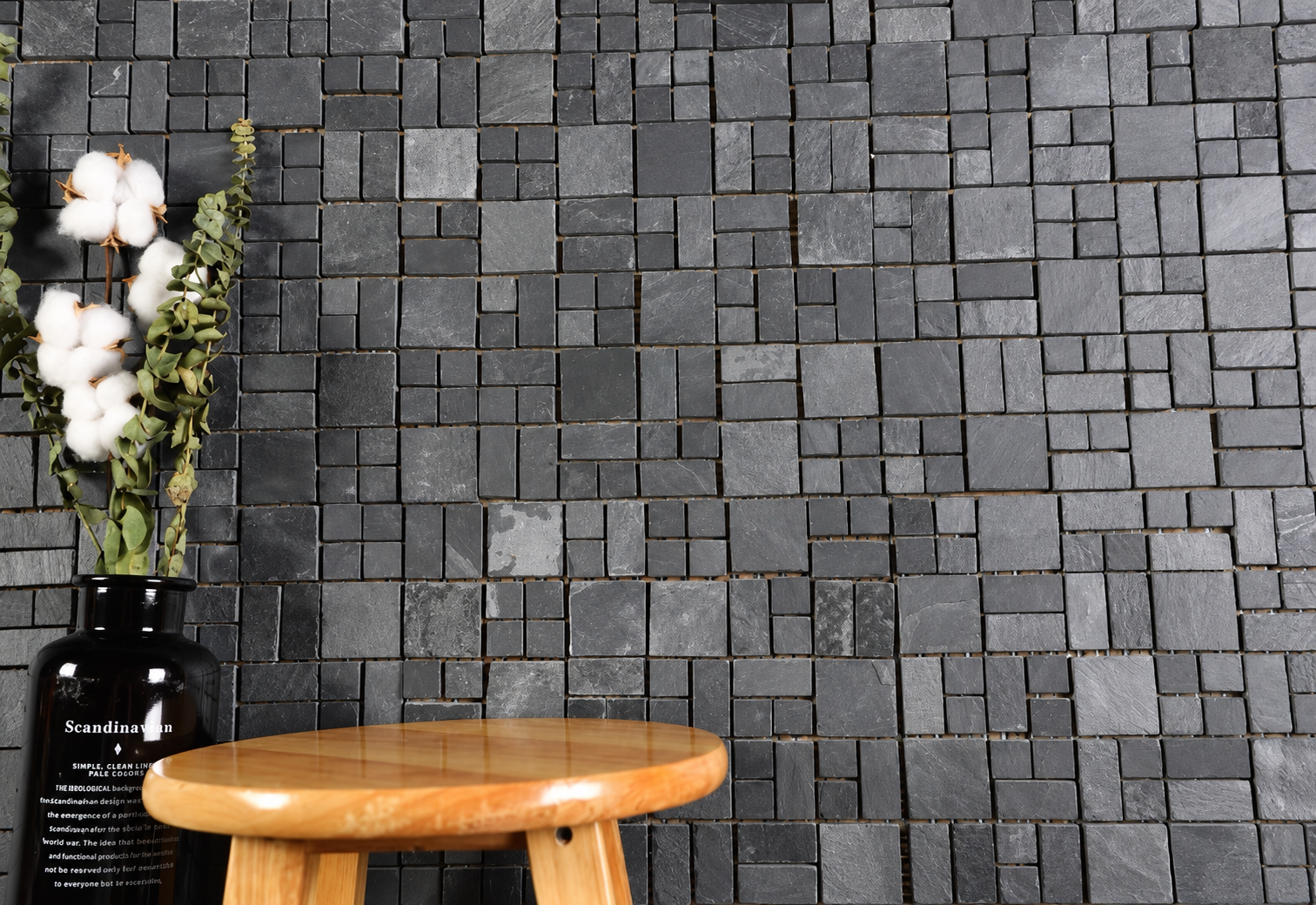 Dark charcoal gray rectangular stone mosaic tile wall with a small round wooden stool and a black vase with cotton stems in the foreground.
