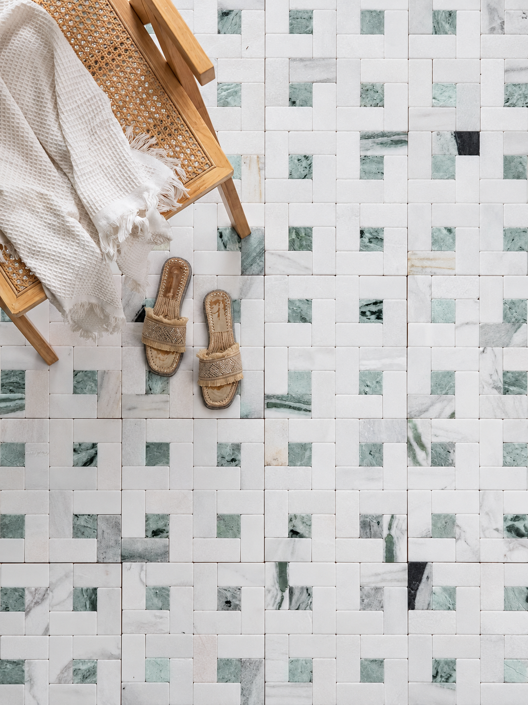 A styled flat-lay featuring a white and green marble mosaic tile floor, with a wooden cane chair draped in a cream knit blanket and a pair of woven jute slide sandals.