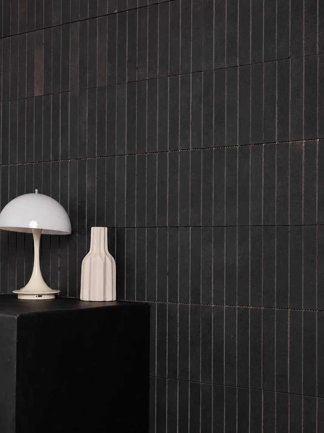 A modern interior scene with a black pedestal holding a white dome lamp and a ribbed white vase, set against a dark charcoal wall with vertical wood slats.