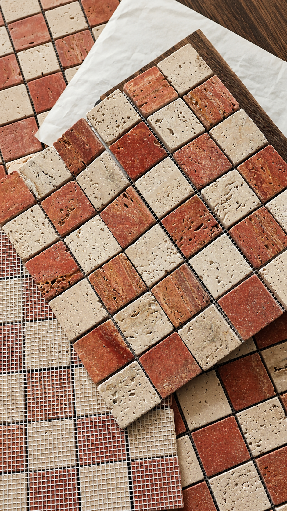 A close-up of mosaic tile samples arranged on a surface, showing square tiles in warm terracotta red, beige stone, and brown tones, with a white sheet partially covering one sample.