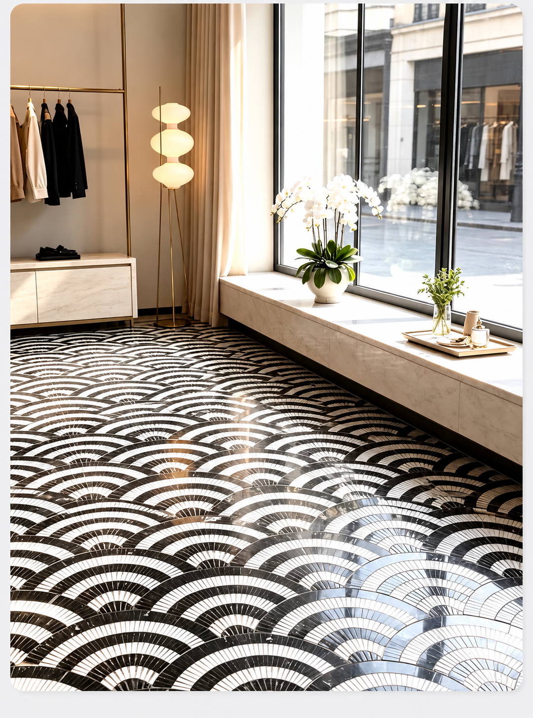 A modern interior space with a large black-and-white geometric fan-patterned floor covering, a long window seat with white flowers, a floor lamp, and a clothing rack in the background.