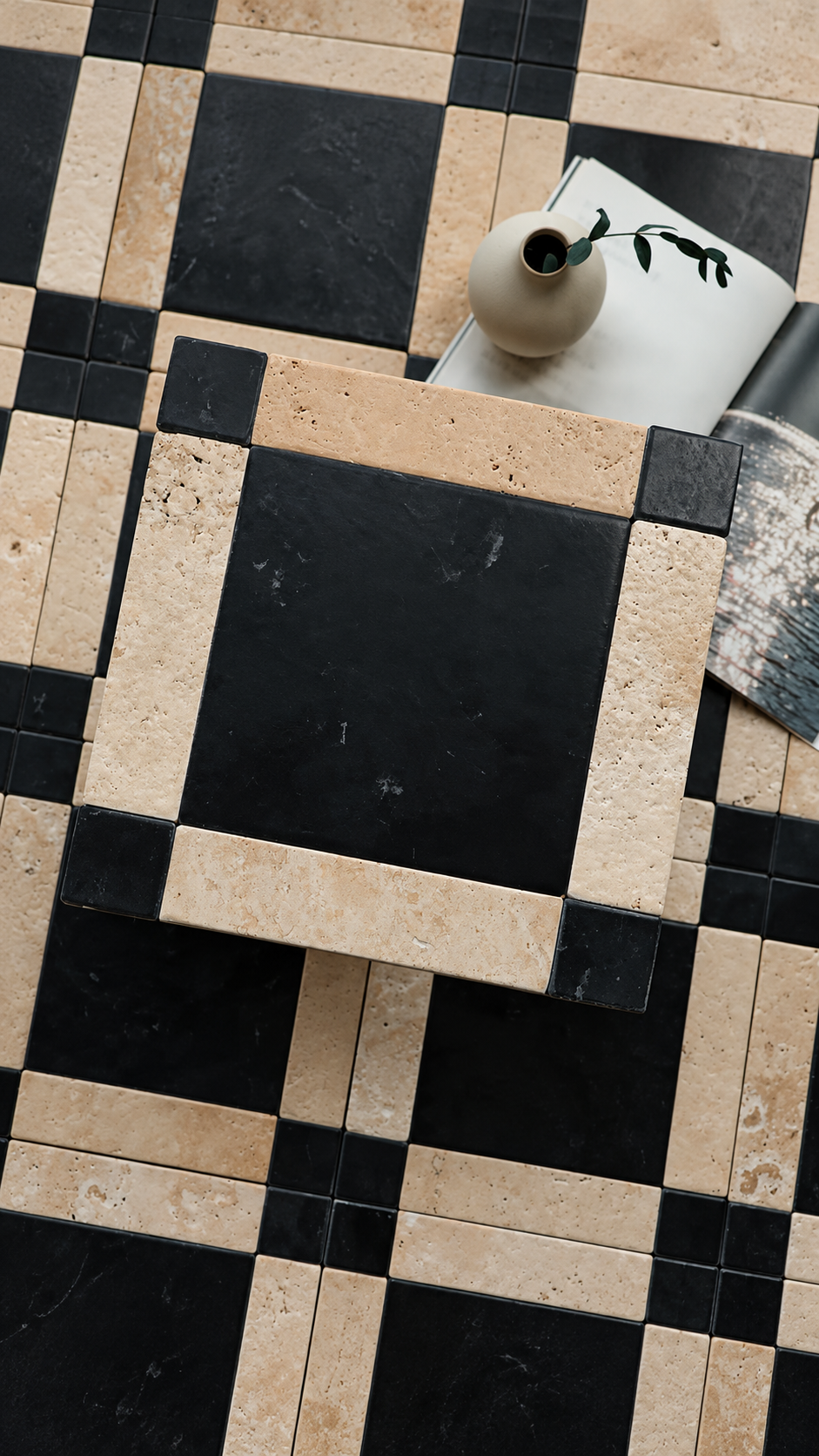 A top-down view of a black and beige checkered tile floor, with a small beige ceramic vase and an open book placed on the surface.