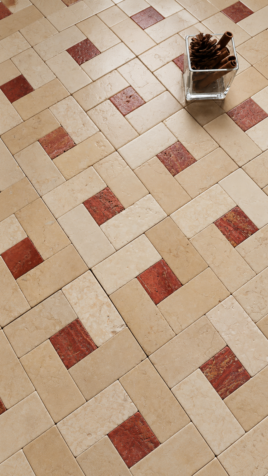 A top-down view of a geometric tile floor pattern featuring beige travertine and reddish-brown stone tiles, with a small glass vase containing pinecones in the upper right corner.