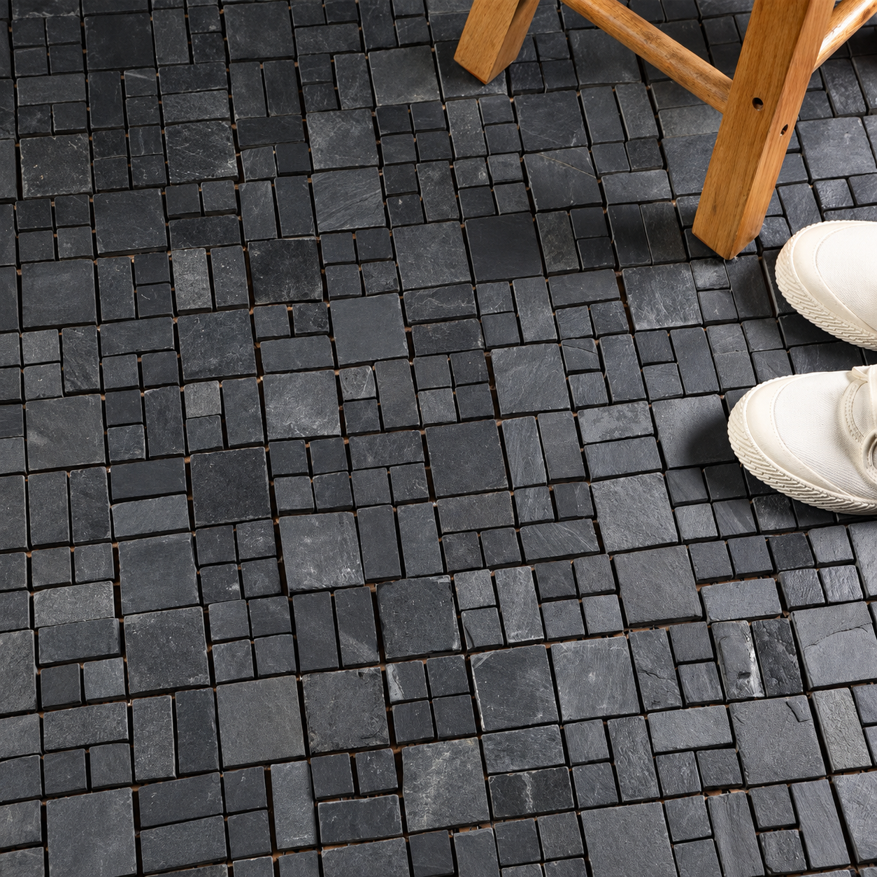 Dark charcoal gray slate mosaic floor tiles with a small square grid pattern, shown in a lifestyle setting with a wooden stool leg and a person's white sneakers visible in the top right corner.