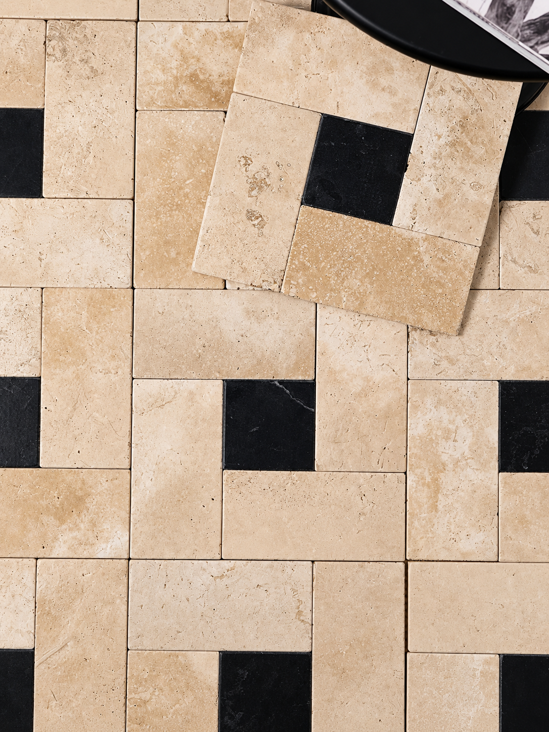 Beige travertine rectangular tiles arranged in a geometric pattern with alternating black square accents, shown with one tile lifted to display its natural stone texture.