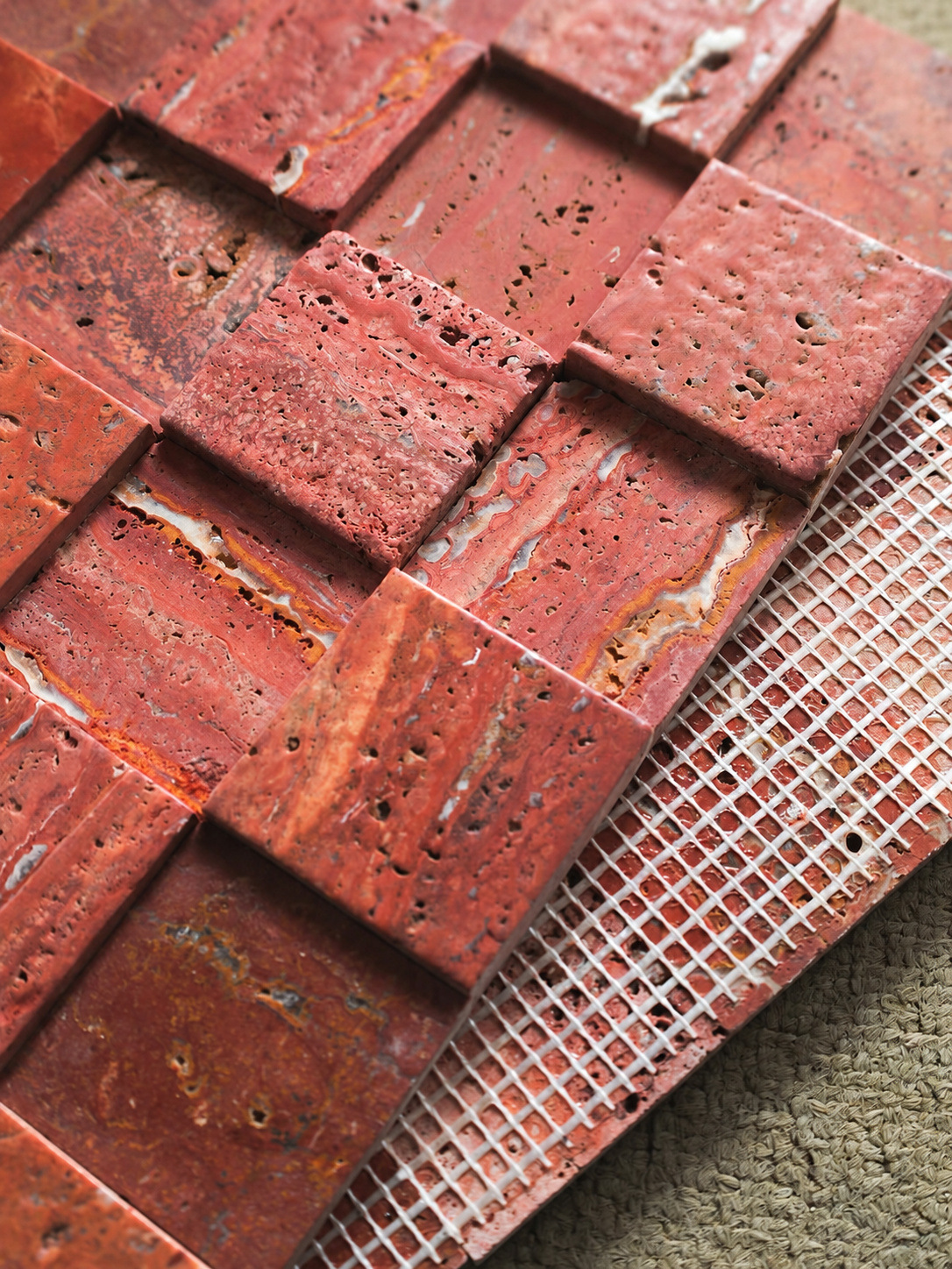 Red textured stone mosaic tiles arranged in a grid pattern, with a white grid overlay showing the backing or mesh layer, photographed close-up on a neutral surface.