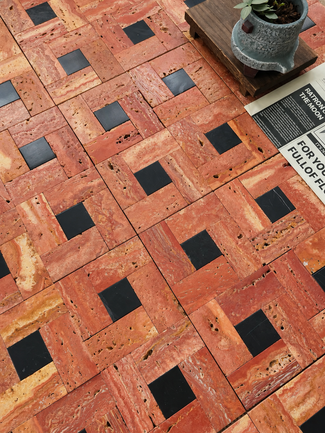Terracotta and orange-brown mosaic tile flooring with alternating black square inlays, shown in a styled interior setting with a small potted plant and a magazine corner visible.