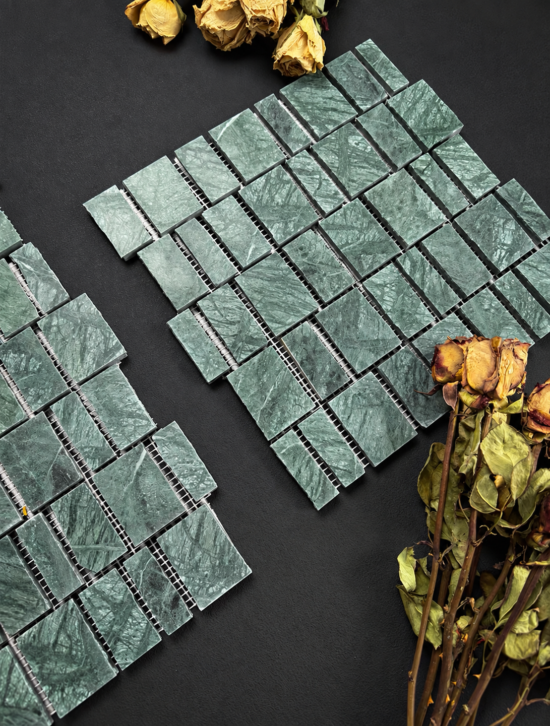 Two rectangular mosaic tile sheets with a textured green marble-like pattern, arranged on a dark background with dried yellow roses as decorative props.