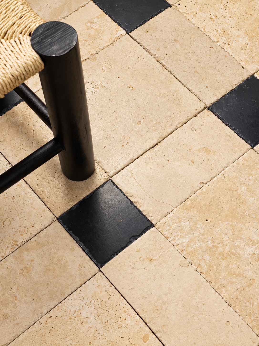 Beige travertine-style square floor tiles with alternating black checkered tiles, shown with the corner of a woven chair and black metal leg visible in the upper left.