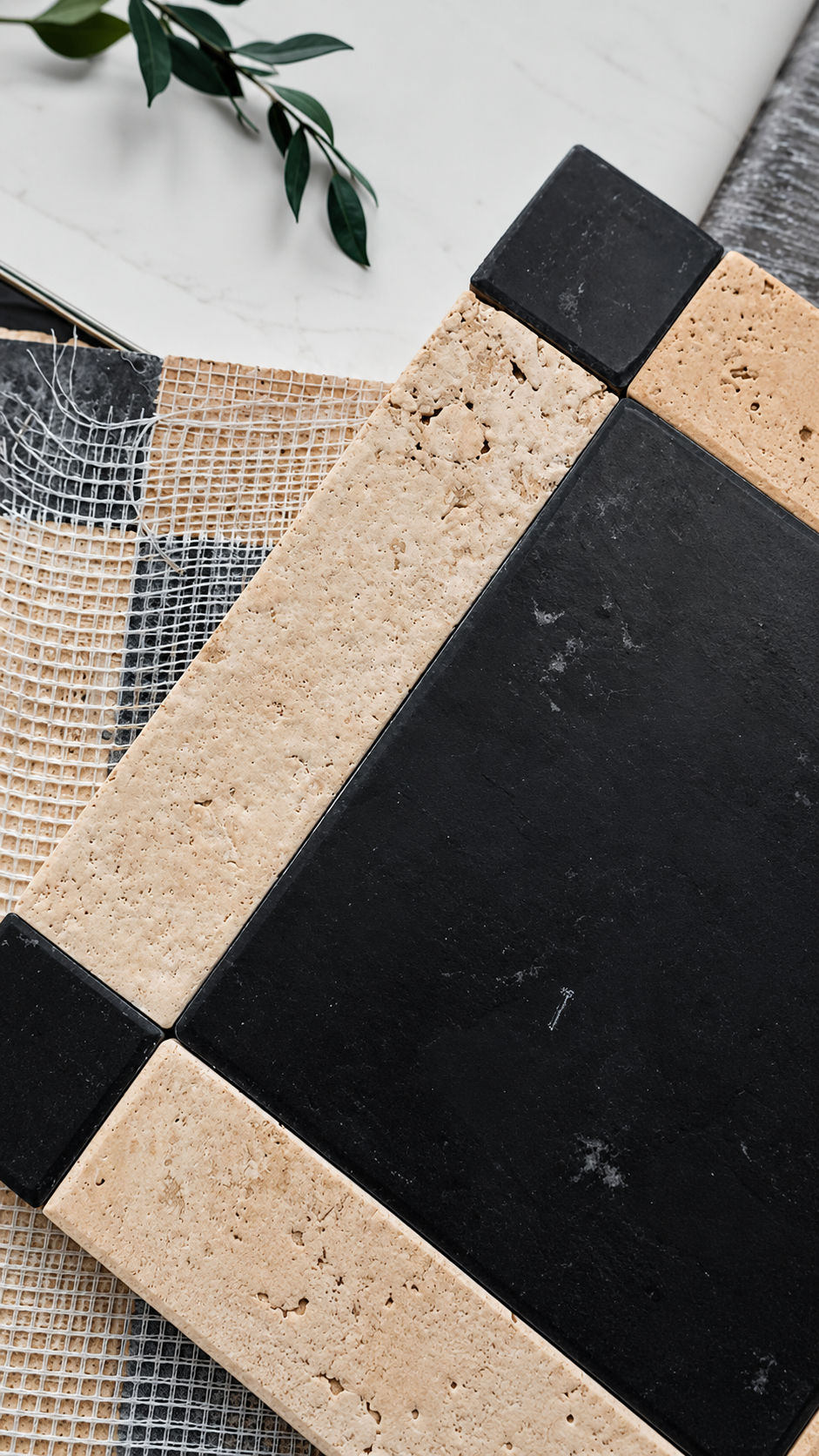 Black and beige stone tile samples arranged diagonally on a white surface with a small green leaf sprig in the corner.