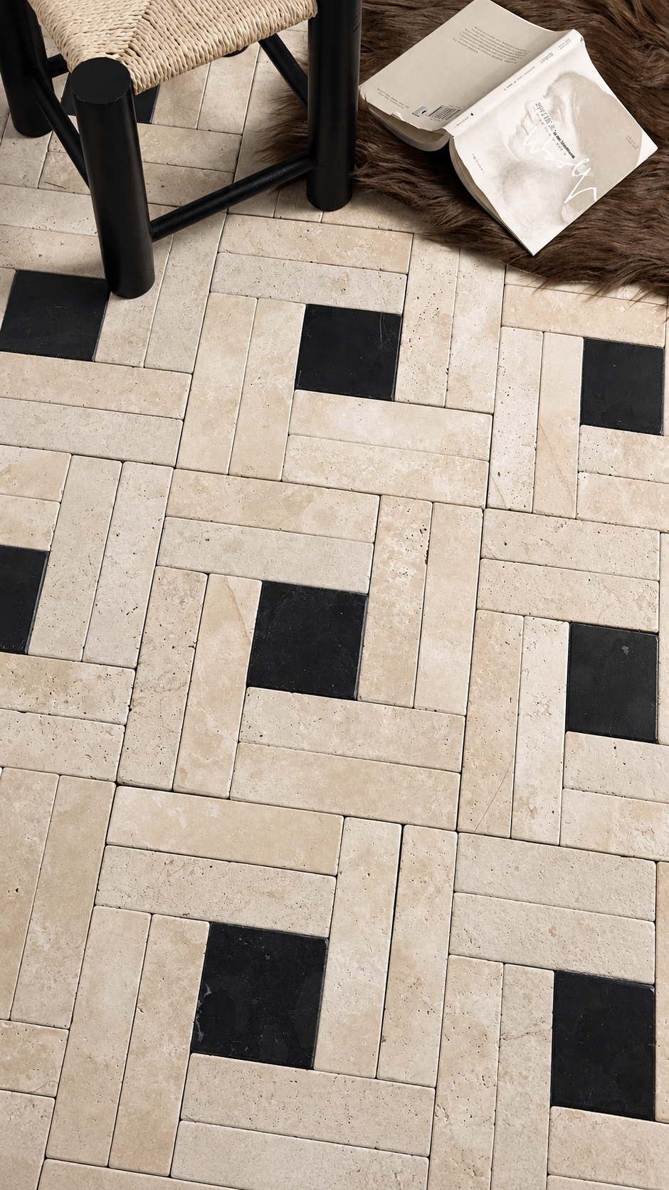 A close-up of a geometric tile floor pattern featuring light beige travertine-style rectangular tiles with alternating black square accents. A woven chair and a white bag are partially visible at the top edge as styling props.
