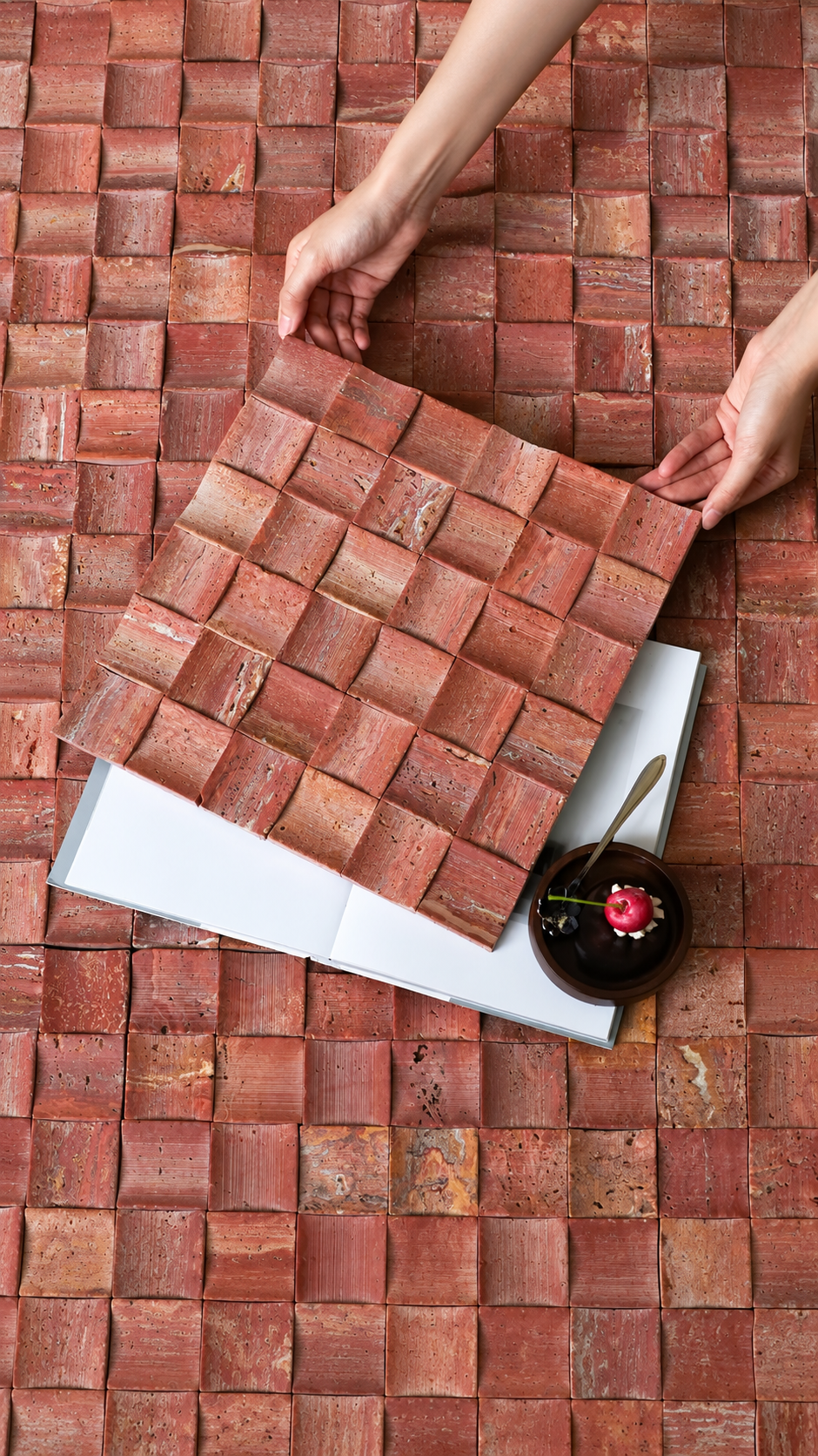 A top-down view of a person's hands holding a square sample of reddish-brown textured tiles over a matching tiled surface, with a white book and a small dark dessert plate nearby.