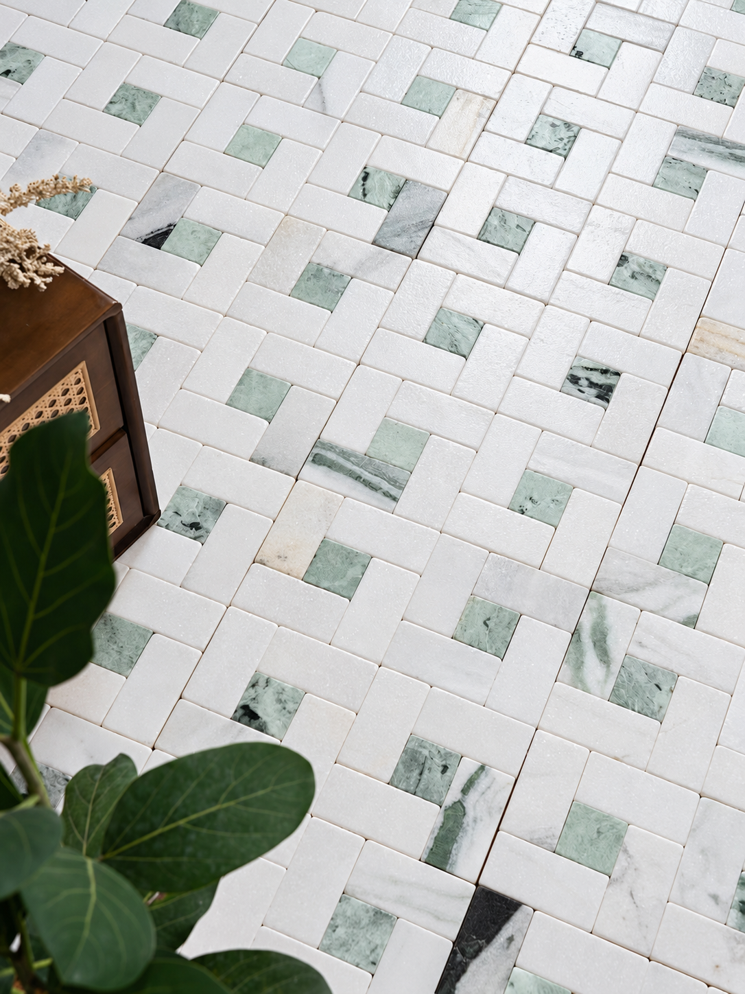 White and light green marble mosaic floor tiles arranged in a basketweave pattern, shown with a dark wood side table and green plant leaves in the foreground.
