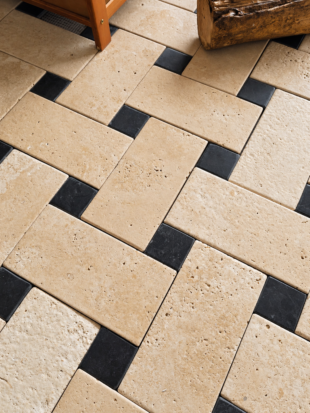 Natural beige travertine stone rectangular tiles arranged in a herringbone pattern with contrasting small black square tiles, shown in a styled interior setting with parts of wooden furniture visible.