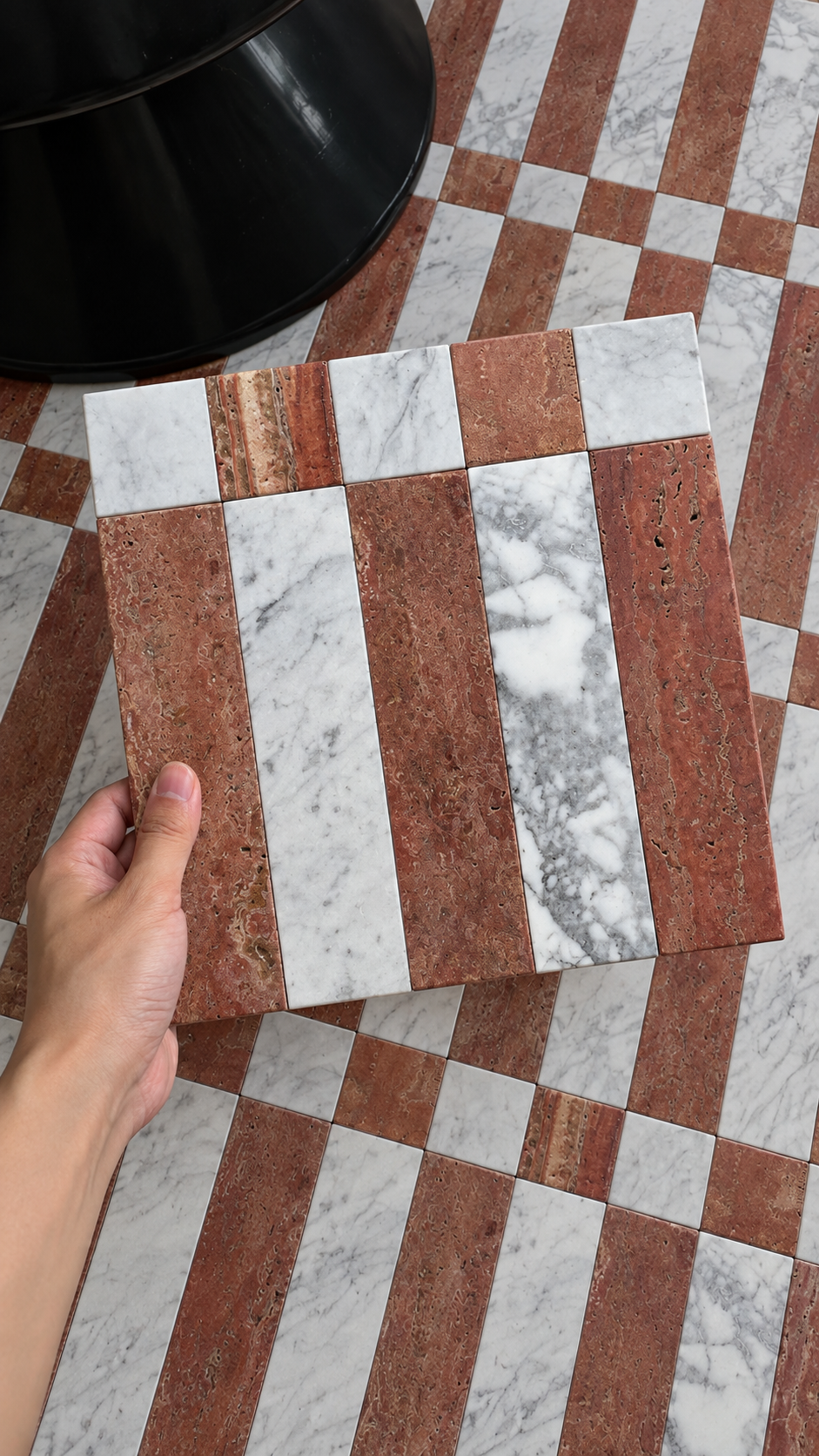 A hand holds a square tile sample featuring alternating vertical stripes of reddish-brown travertine and white marble, set against a larger matching checkered floor pattern.