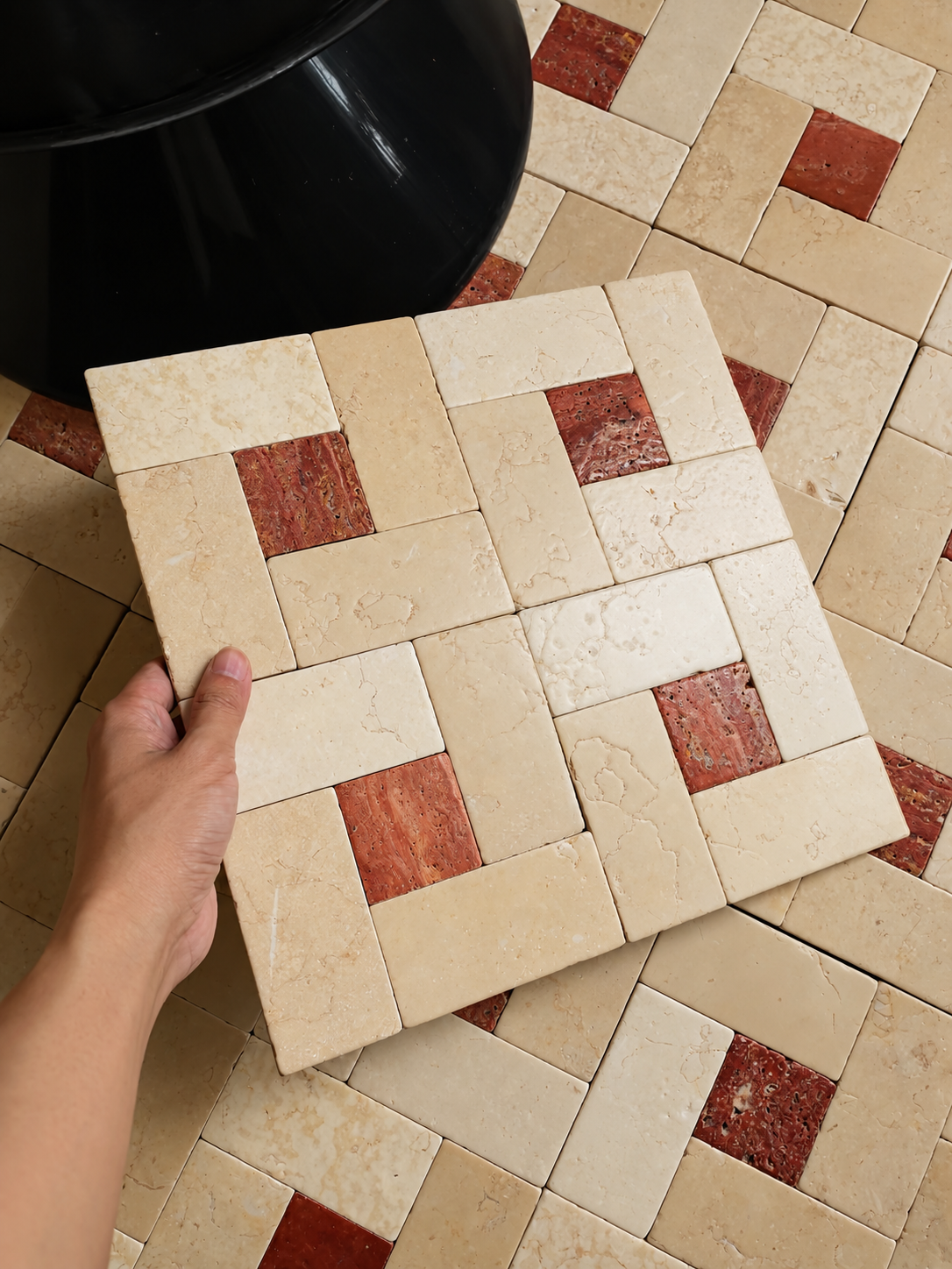 A hand holds a square tile sample showing a beige and reddish-brown mosaic pattern, with a larger matching tile sample laid out on the floor below.