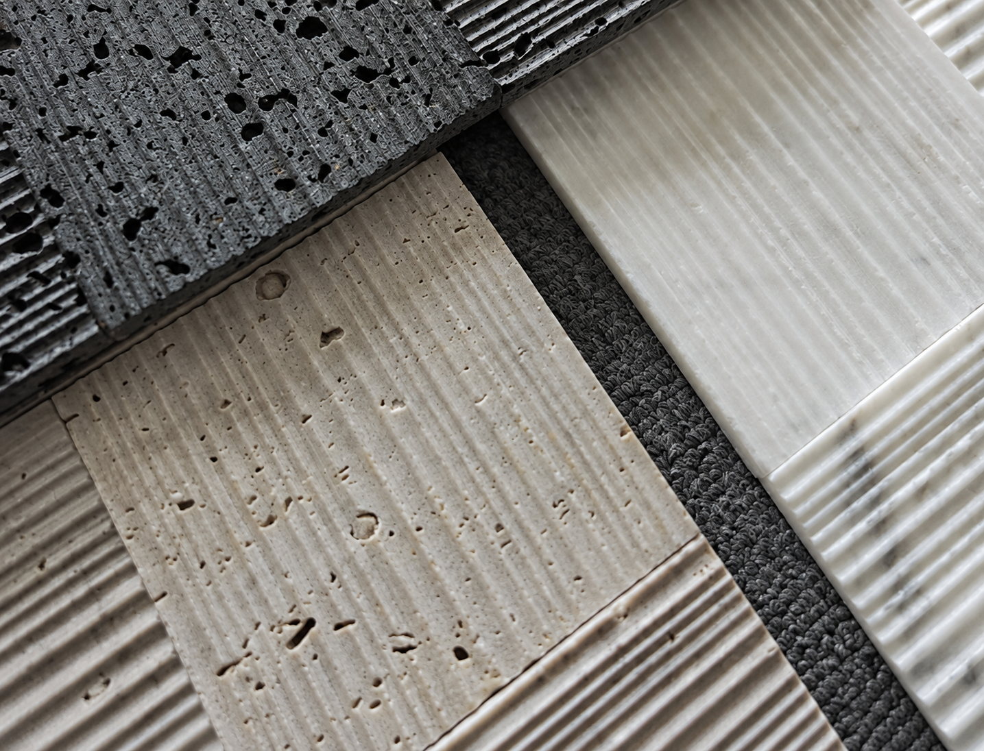 Close-up of textured stone tile samples arranged diagonally, showing dark charcoal and warm beige surfaces with ribbed and porous finishes.