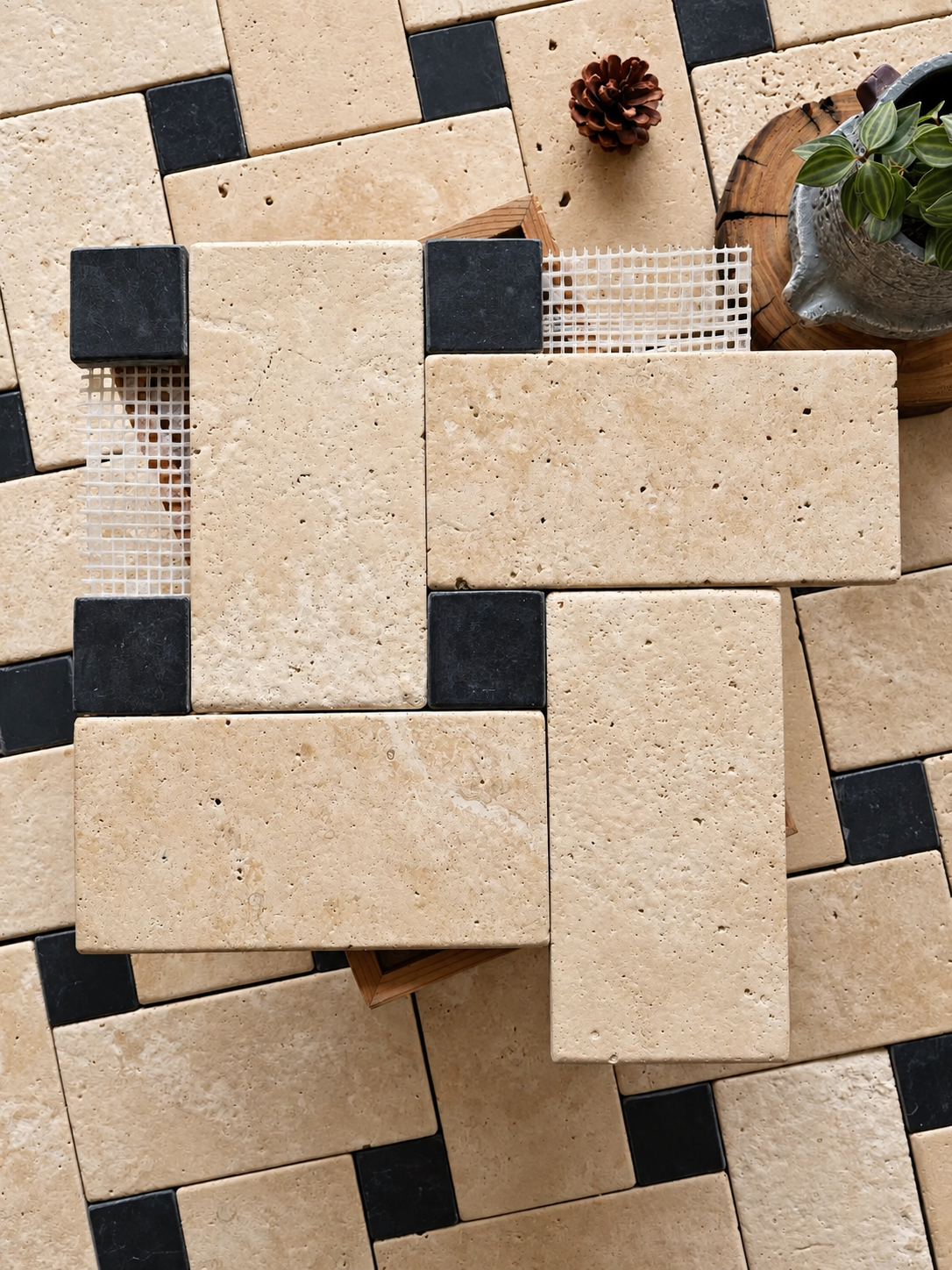 Natural beige travertine stone rectangular tiles arranged in a geometric pattern with black accent tiles, shown in a styled flat-lay composition with a small potted plant and pinecone.