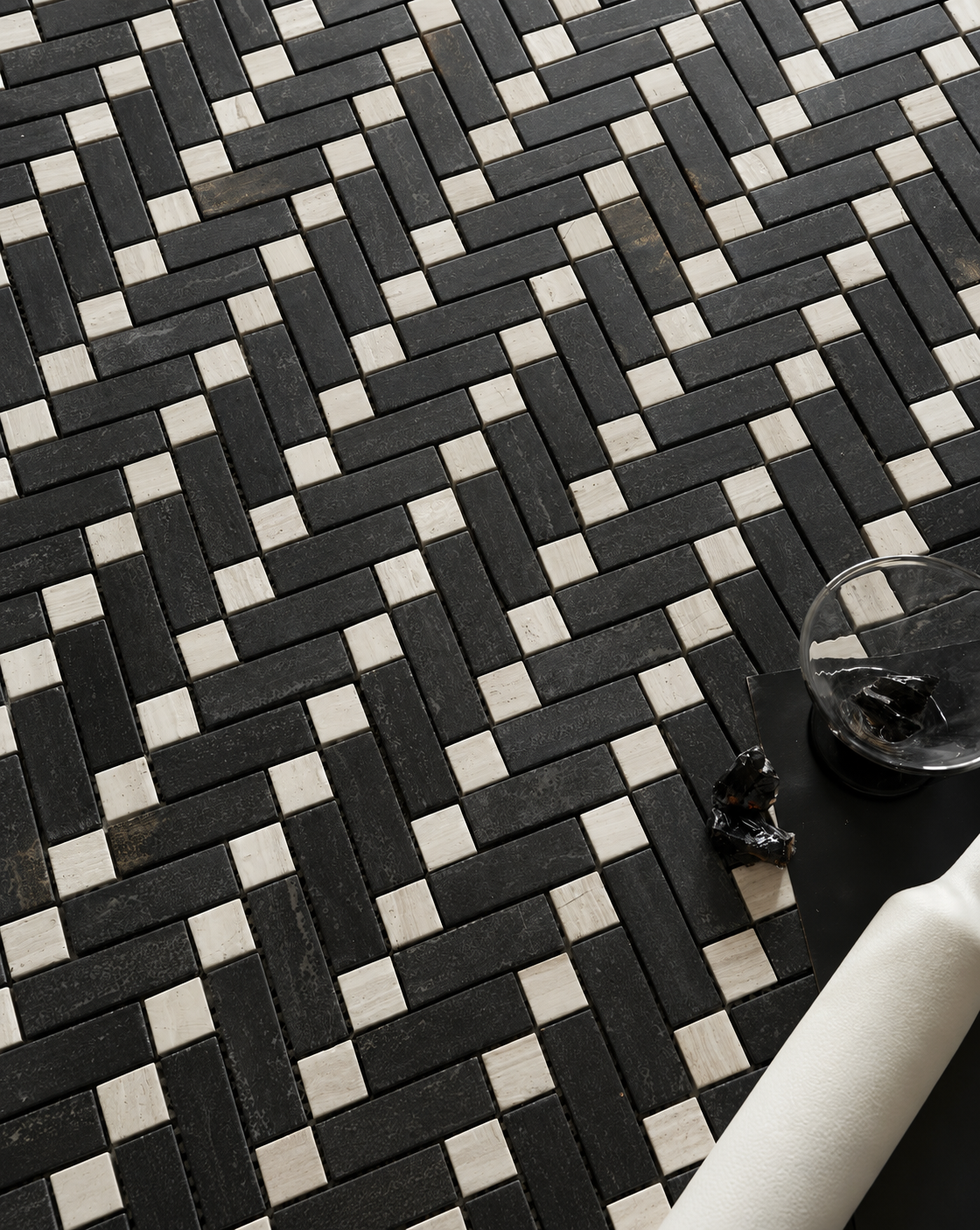 Black and white woven herringbone mosaic tile floor with a small glass bowl and dark decorative object on a dark surface in the lower right corner.