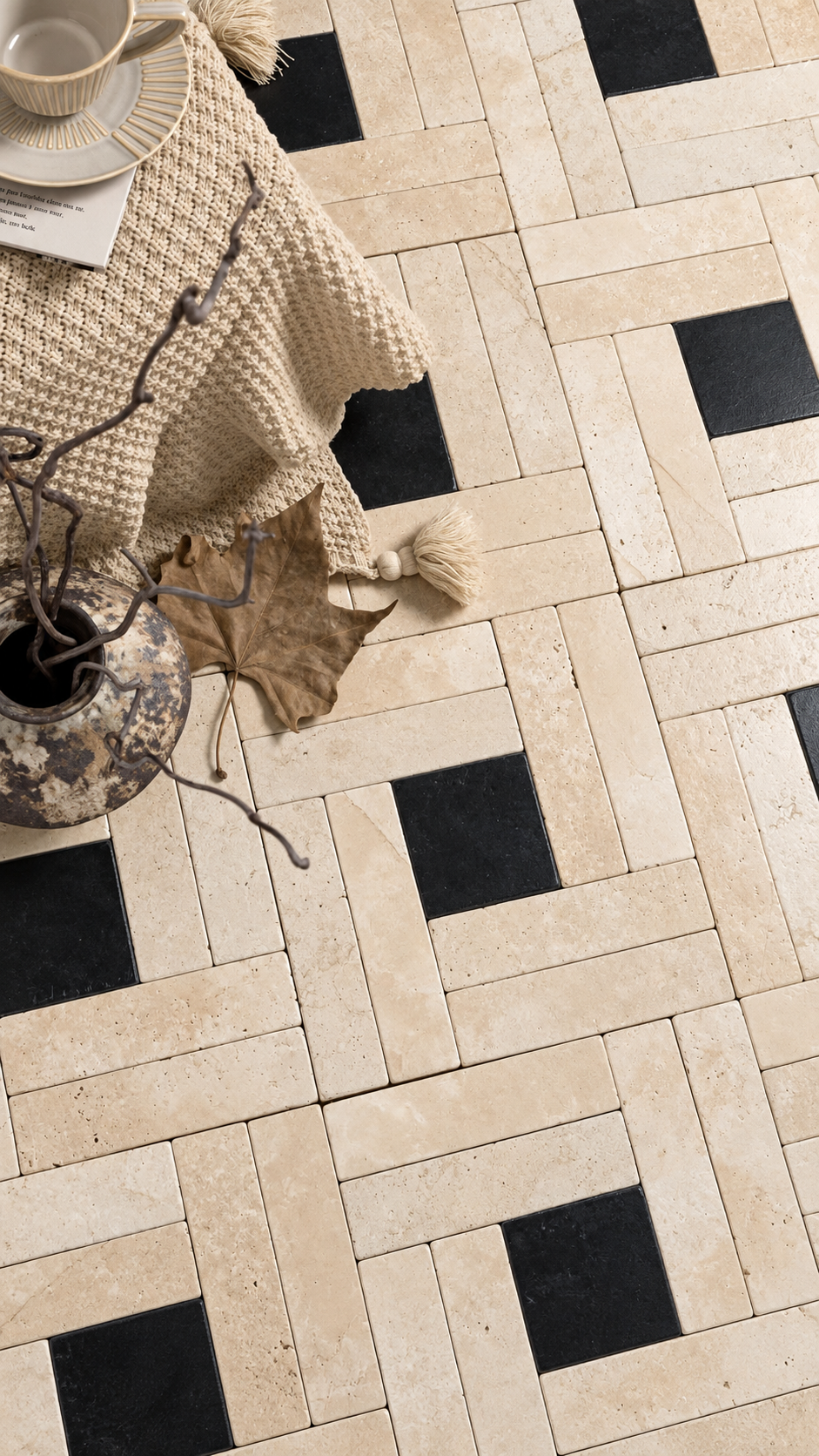 Beige travertine-style rectangular floor tiles arranged in a herringbone pattern with alternating black square accents, styled with a cream tablecloth, ceramic vase, dried leaves, and a cup in the upper left.