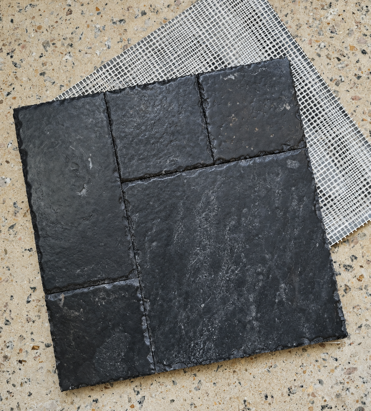 A square black slate tile sample with a rough natural stone texture, shown on top of a white mesh backing over a speckled beige surface.