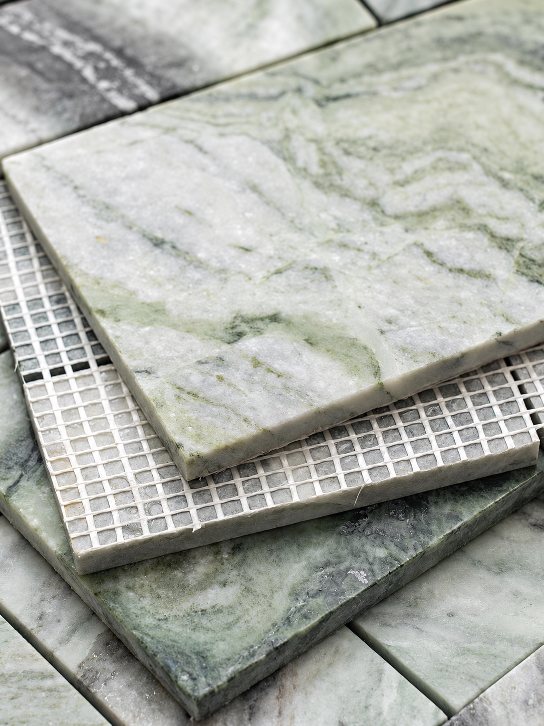 Close-up of stacked square marble tiles in pale green and gray tones, with one large tile resting on a smaller tile and a grid-patterned tile sample underneath.