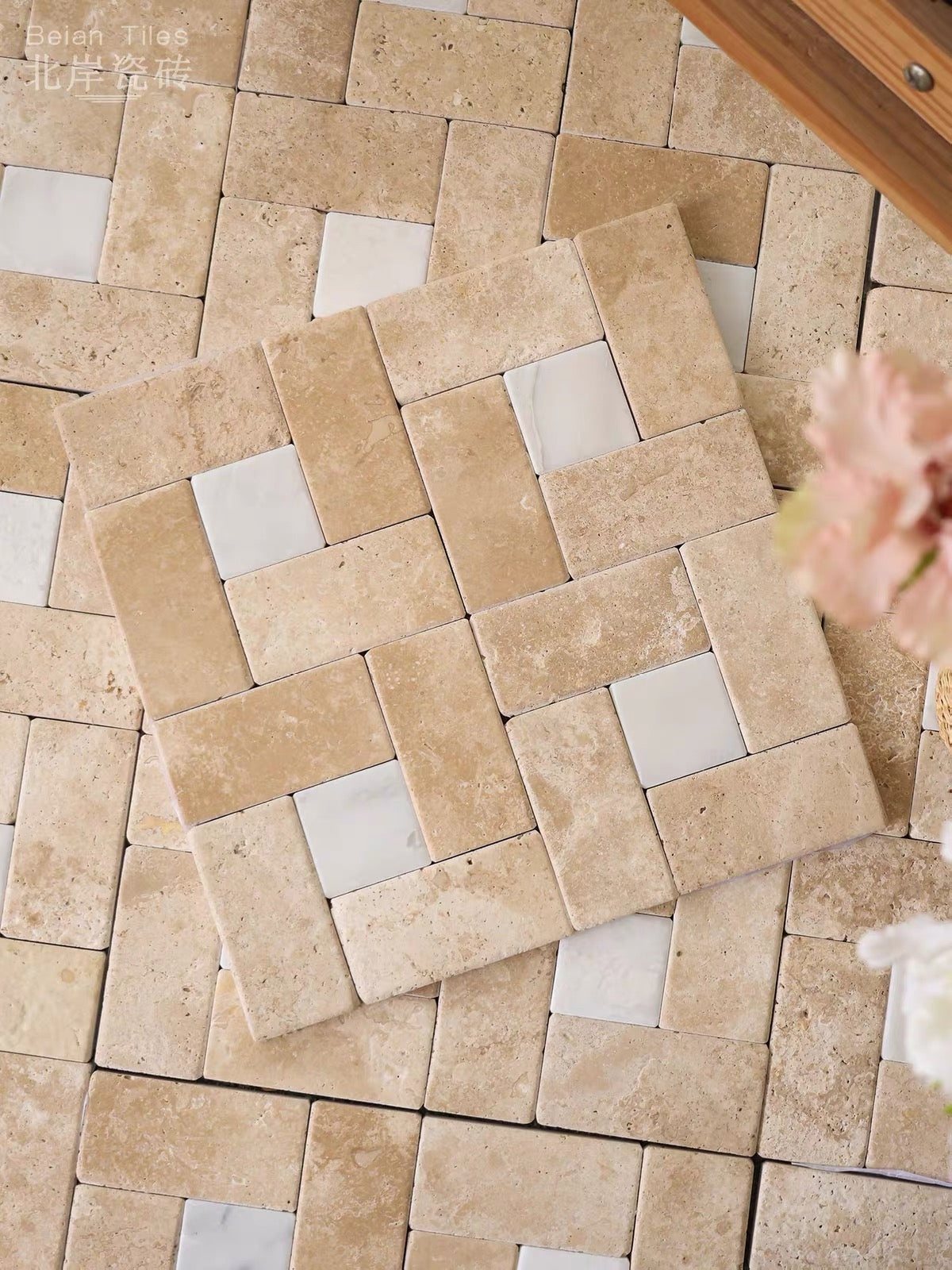A square mosaic tile sample with a basketweave pattern of beige travertine-like stone and white marble accents, displayed on top of a matching full tile sheet. The background features a wooden surface and soft pink flowers in the corner.