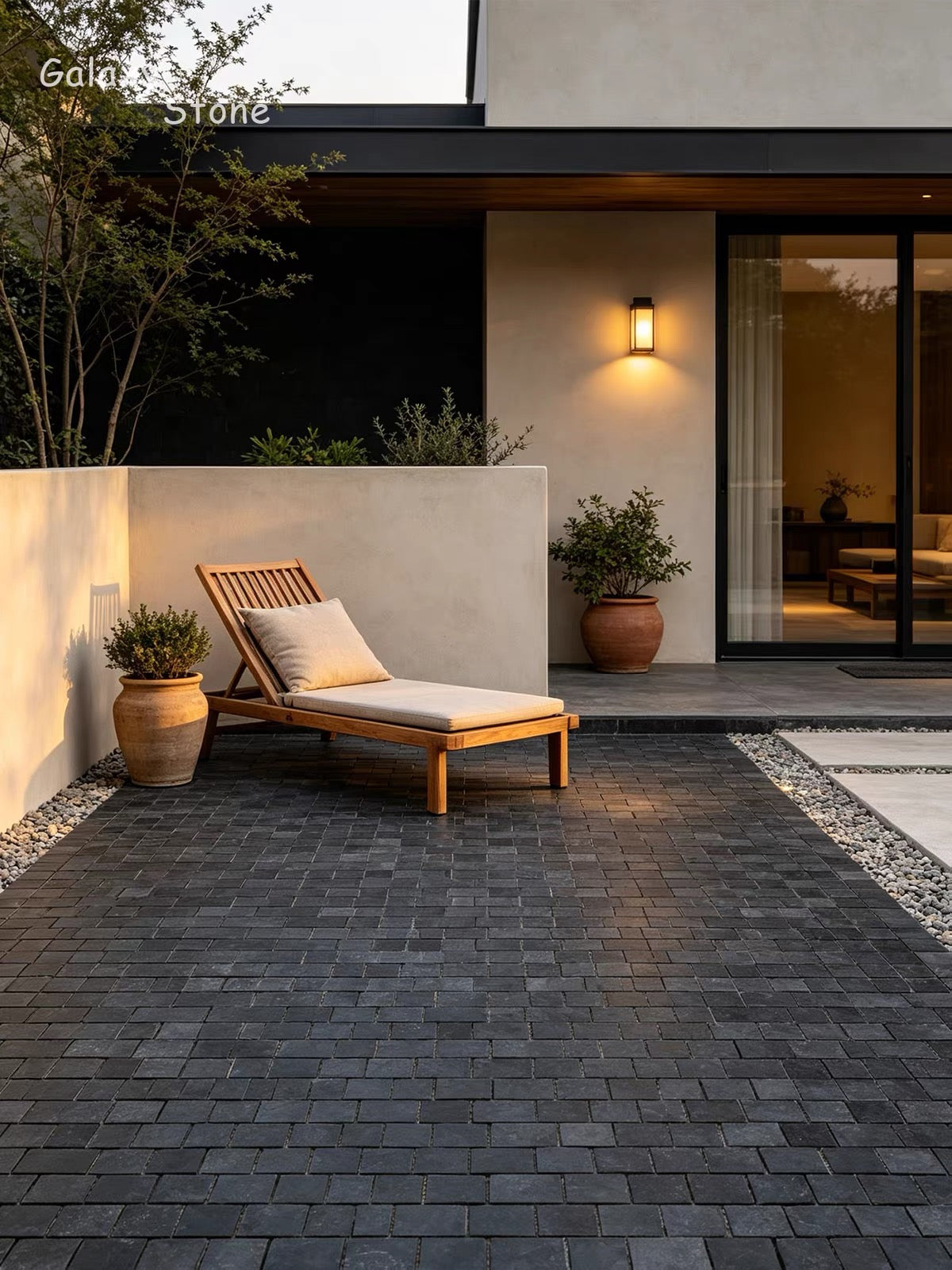Dark gray rectangular stone pavers arranged in a modern outdoor patio setting next to a wooden lounge chair with a cream cushion, potted plants, and a contemporary house facade.