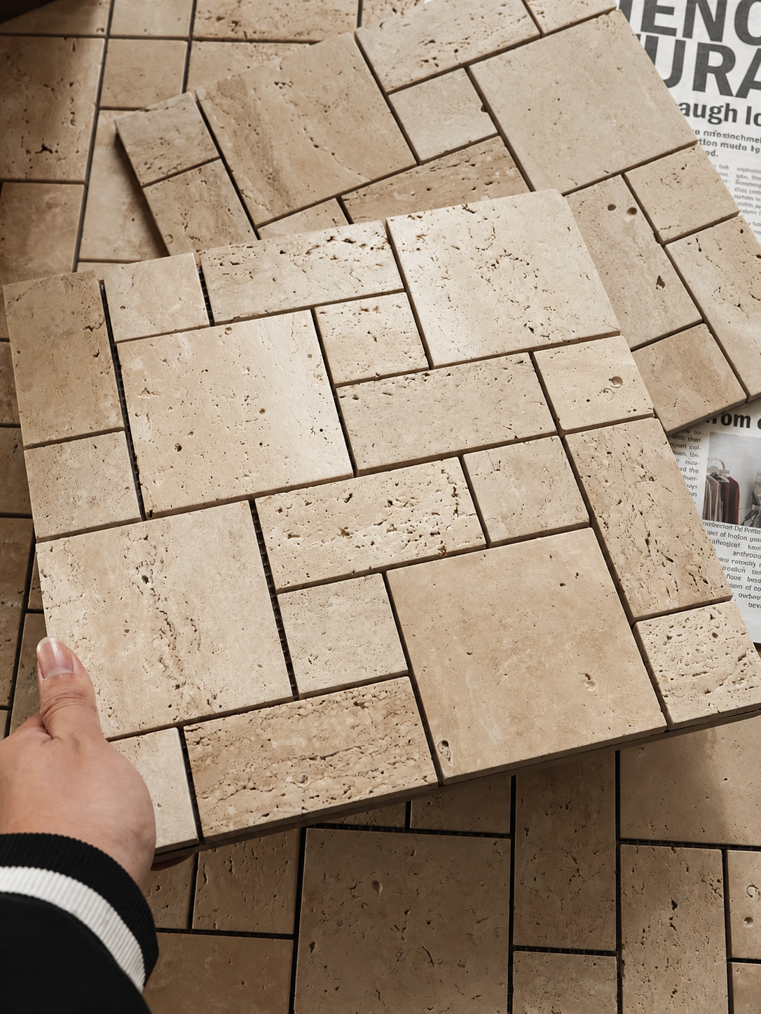 A hand holds a square tile sample showing a beige travertine-style stone pattern with dark grout lines, with additional matching tile samples and a newspaper visible in the background.