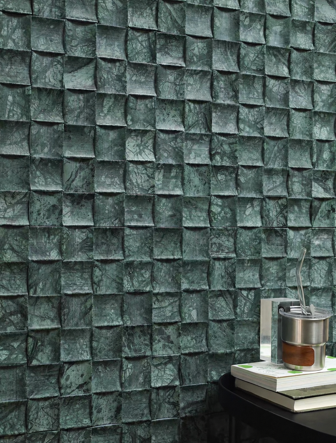 Dark green textured wall tiles with a 3D geometric cube pattern, shown in a styled interior setting with a small table and books in the lower right corner.