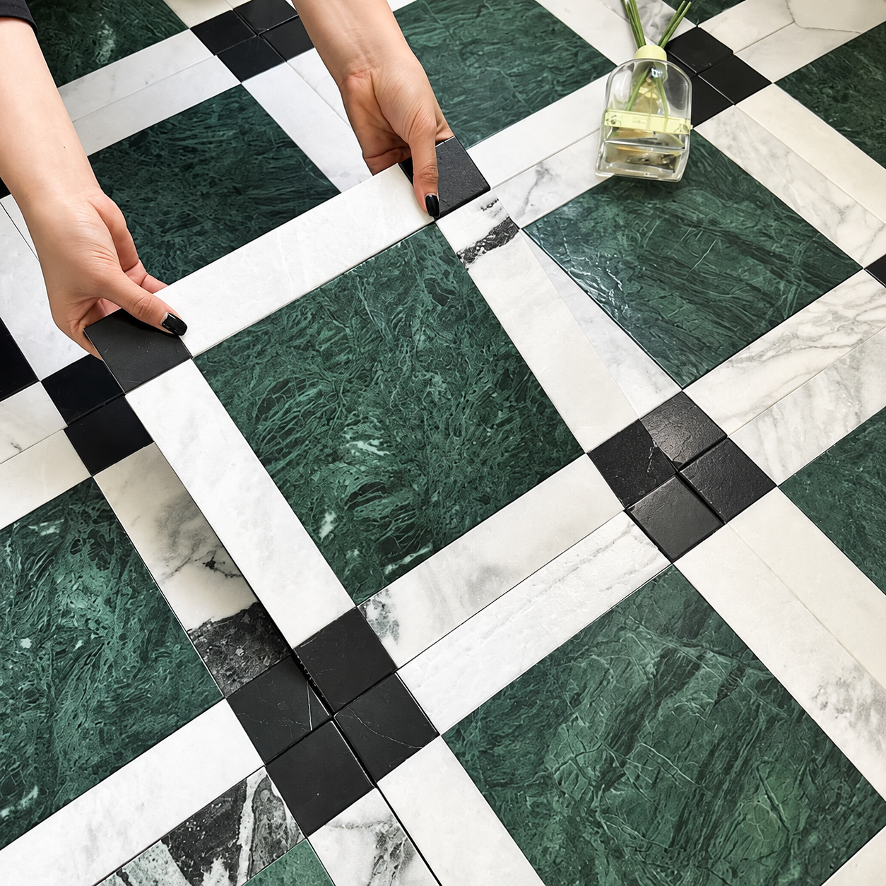 A close-up of a checkered floor made of square tiles, featuring dark green marble-look tiles, white marble-look tiles, and black accent tiles. Two hands are shown holding a black tile, and a small glass vase with green stems sits in the upper right corner.