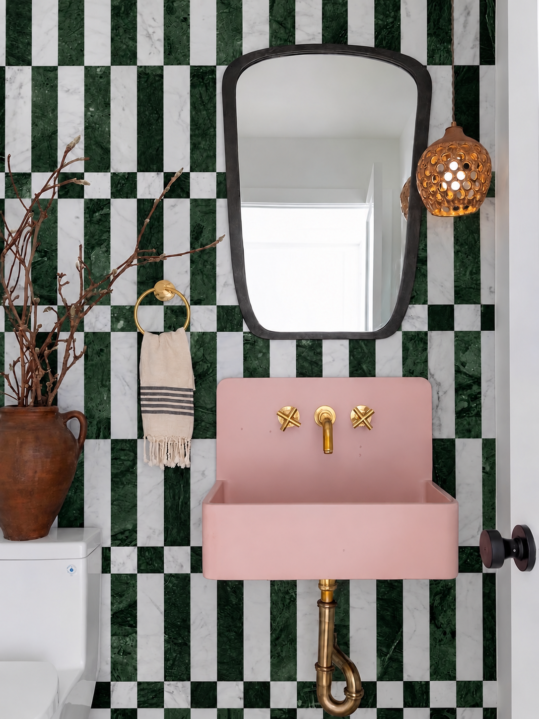 A pink rectangular wall-mounted sink with gold faucet hardware, set against a green and white checkered tile wall. A black-framed mirror hangs above, with a woven towel on a gold ring holder to the left, and a warm woven pendant light on the right.