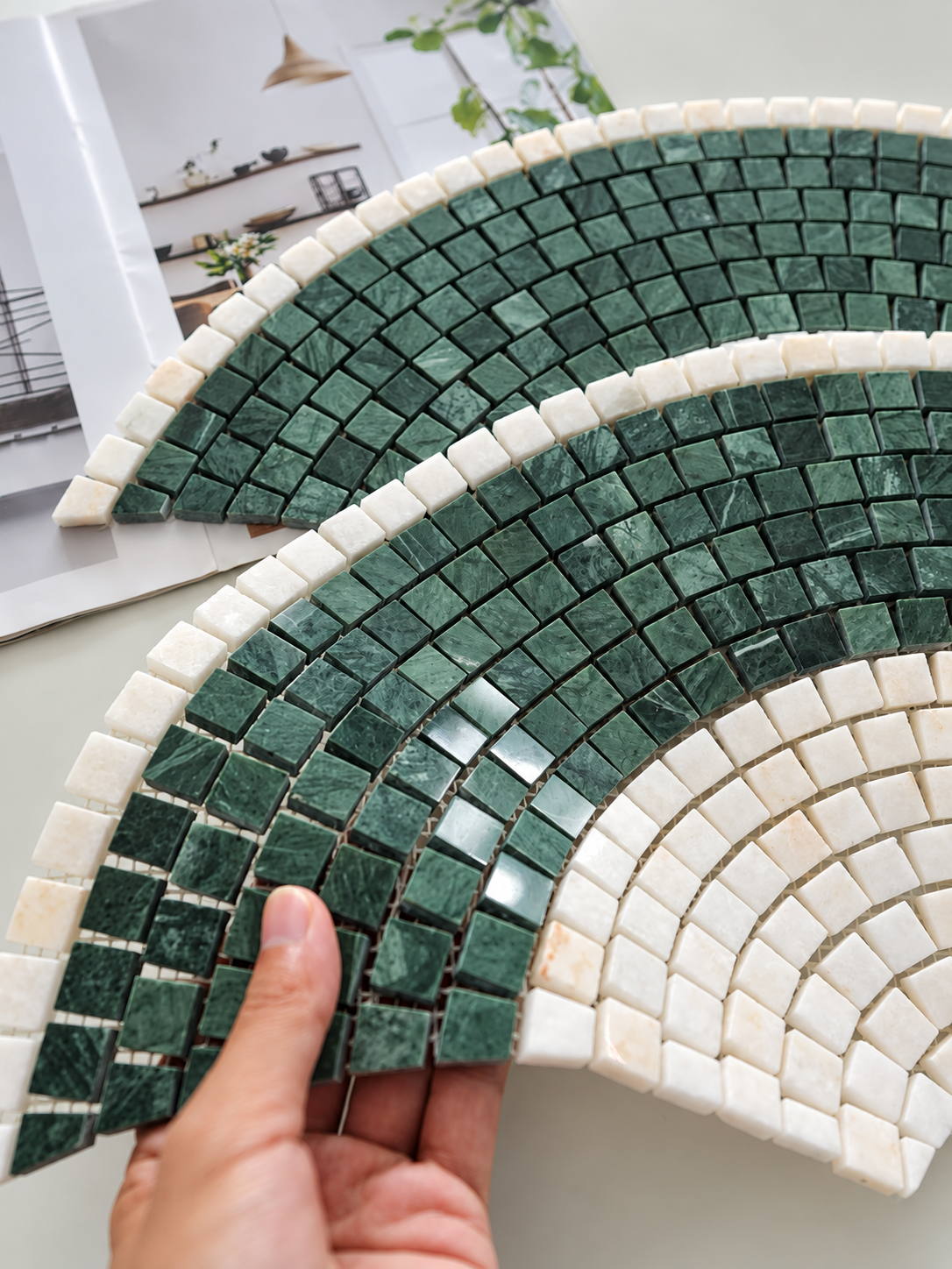 A hand holds two semi-circular mosaic tile samples featuring green marble-like tiles and white marble-like tiles, with a magazine page visible in the background.