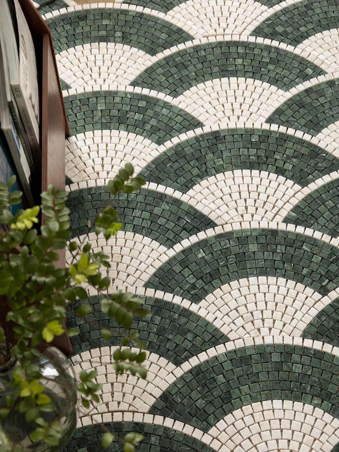 Green and white geometric mosaic tile pattern with repeating scallop or fan shapes, shown in a close-up interior setting with part of a wooden frame and greenery visible at the left edge.