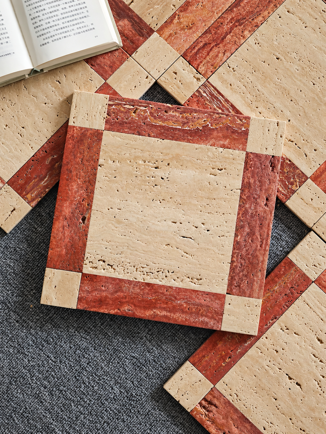 Square travertine stone coasters arranged in a geometric pattern on a dark gray textured surface, with an open book partially visible in the top-left corner.