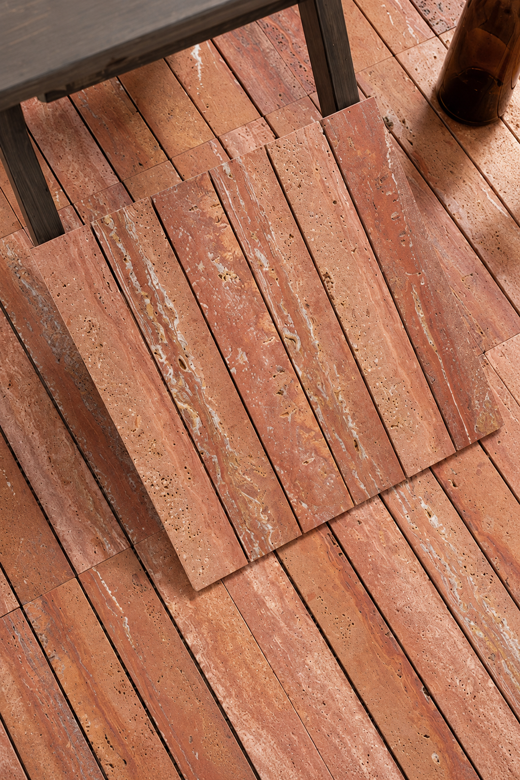 Terracotta-colored travertine stone tiles arranged on a wooden deck floor, with a dark table leg and a brown bottle visible in the scene.