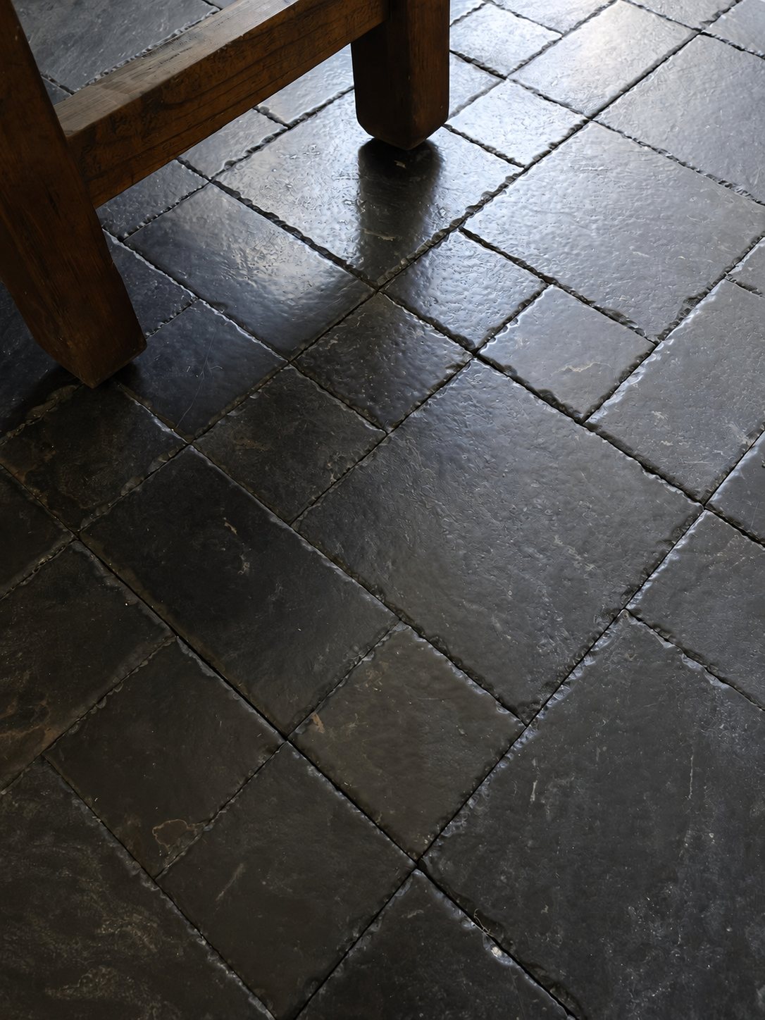 Dark charcoal gray stone floor tiles with a rustic look, shown with the corner of a wooden stool visible in the upper left.