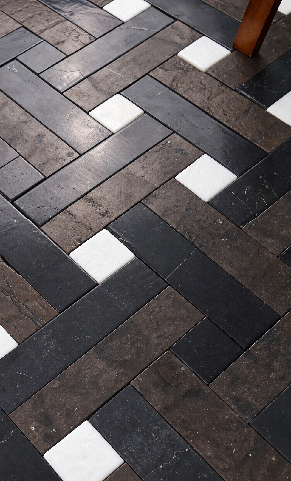 A close-up of a herringbone-patterned floor tile or wood-look plank surface in dark charcoal, brown, and white, with a small portion of a wooden leg visible in the top right corner.