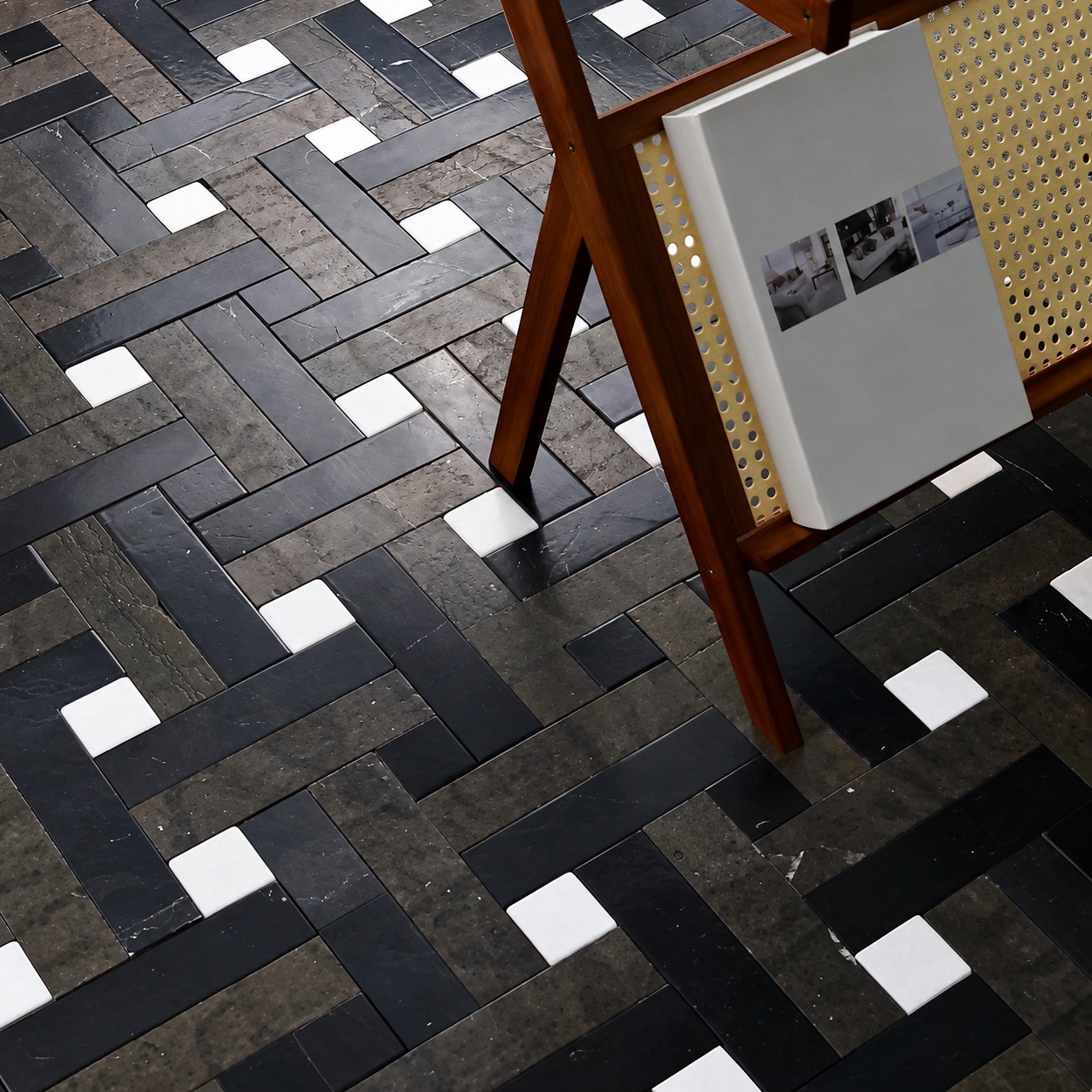 A close-up, high-angle view of a modern geometric floor tile pattern featuring black, dark gray, and white square tiles arranged in a herringbone layout, with a wooden chair leg and a magazine resting on the right side.
