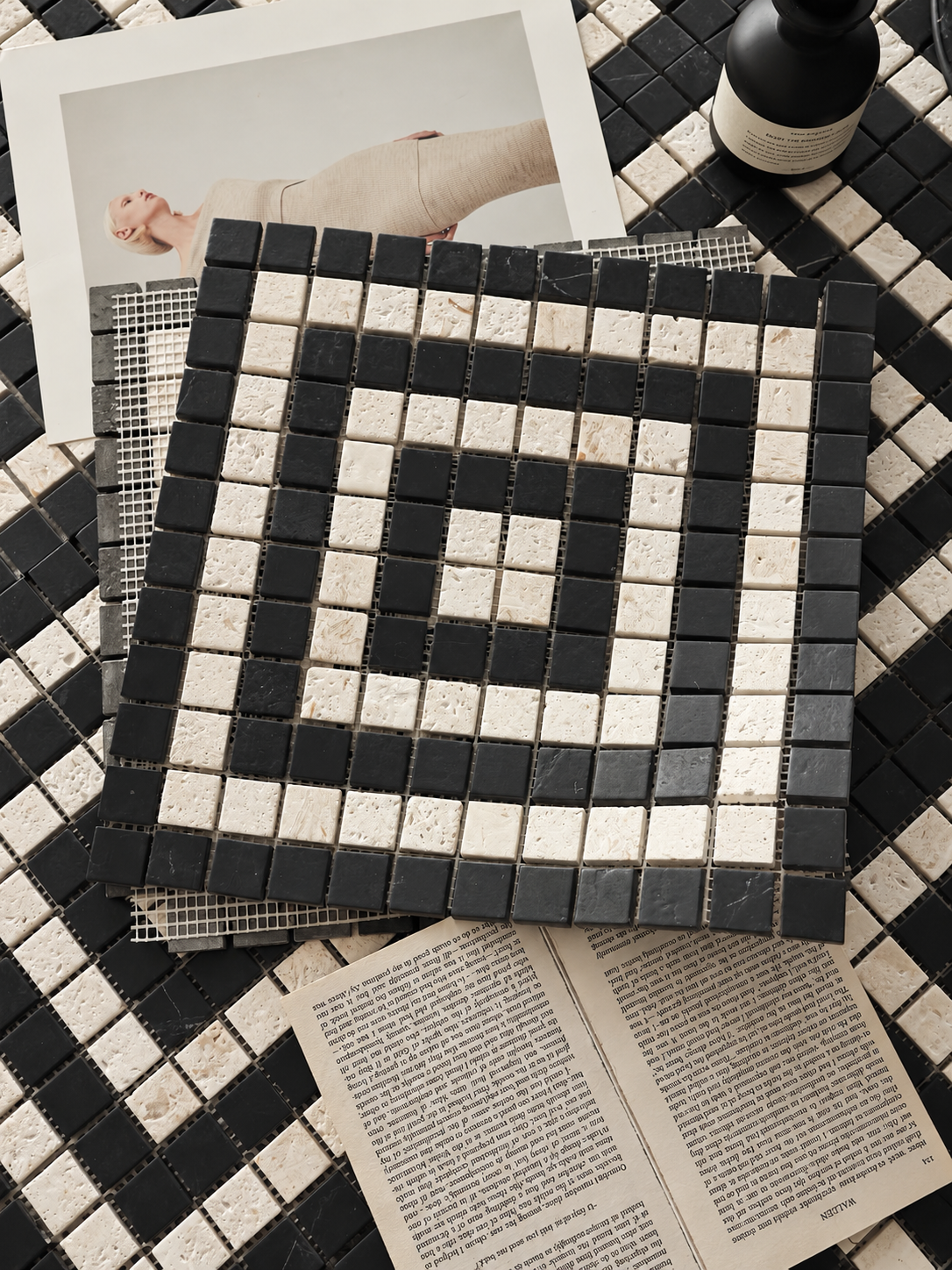 A square mosaic tile sample with a black and off-white geometric pattern, displayed on top of a matching black and off-white tiled surface. The scene includes styled props: an open book, a magazine page with a photo, and a dark bottle.
