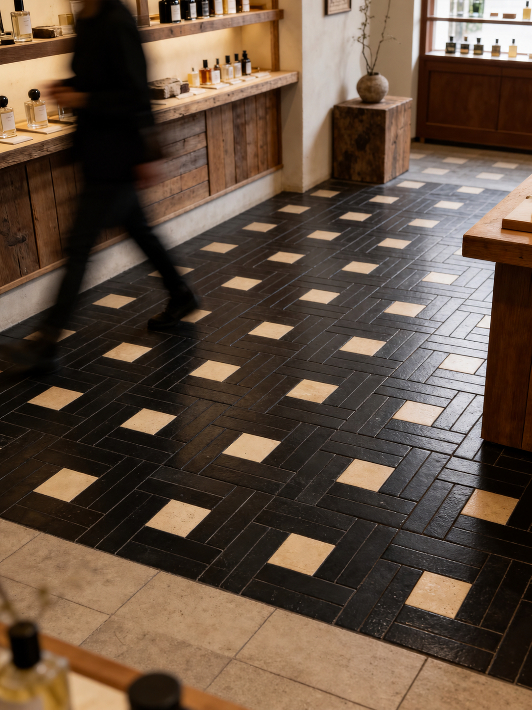 Black and beige geometric patterned floor tiles arranged in a herringbone layout inside a rustic wooden shop interior, with a blurred person walking in the background.
