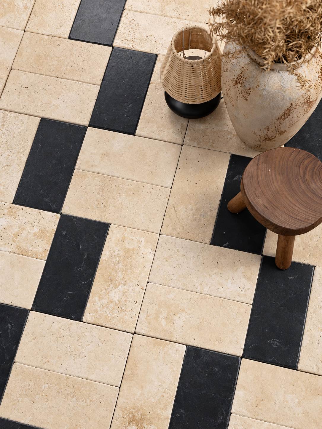 A top-down view of a checkered tile floor featuring alternating beige travertine and black stone tiles, styled with a small wooden stool, a woven basket, and a large ceramic vase with dried grass.