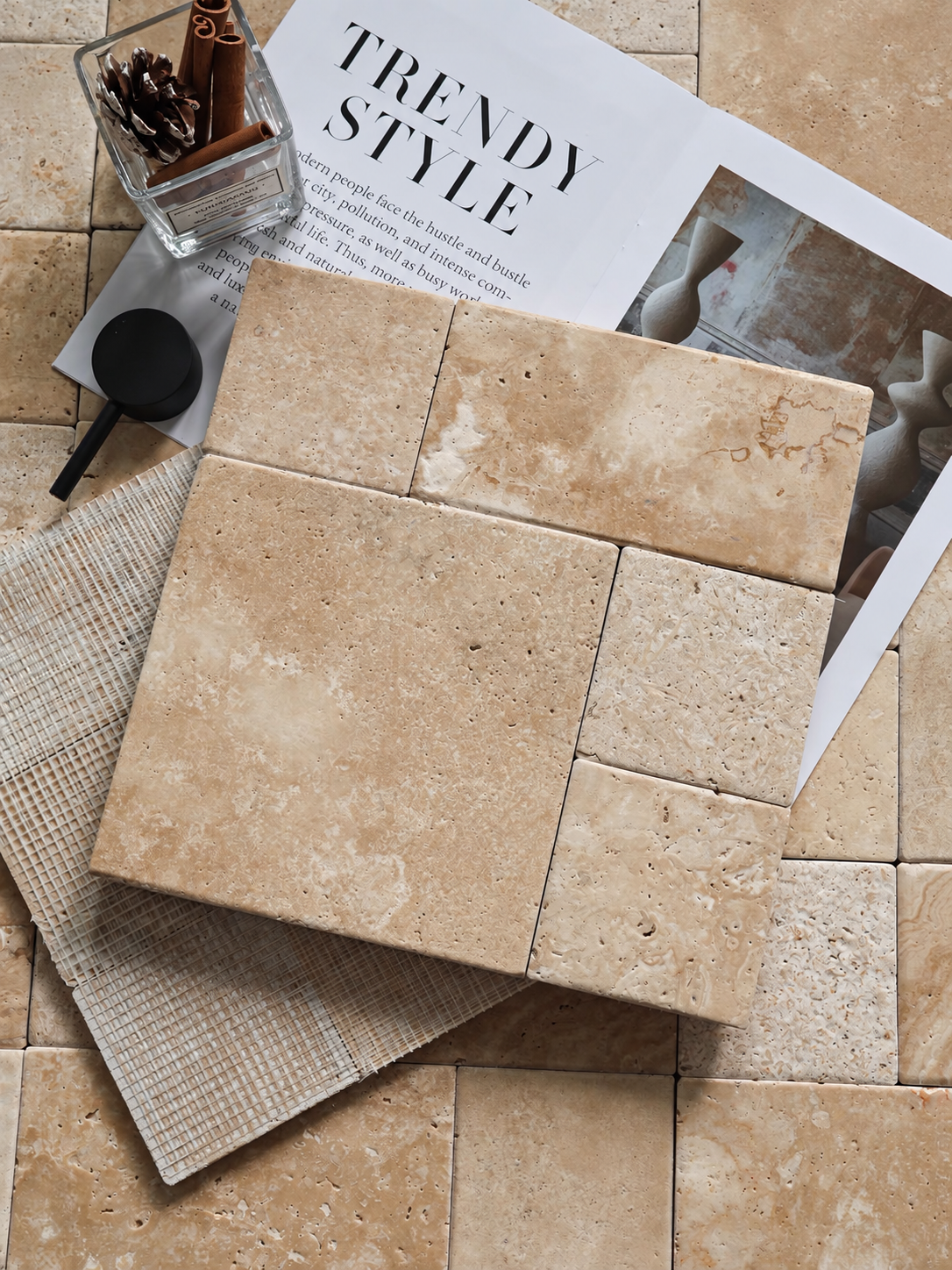 Square travertine-style tiles in a warm beige and cream natural stone finish, arranged on a matching tiled floor with a magazine, small glass vase, and woven mat as styling props.